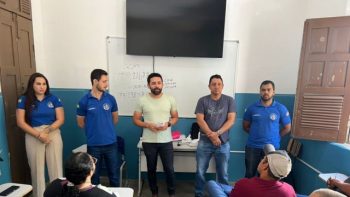 Um grupo de cinco pessoas está em pé diante de uma sala de aula, falando para um pequeno público sentado. Atrás deles há uma lousa branca e uma televisão fixada na parede. Os participantes parecem estar em um momento de apresentação ou reunião institucional.