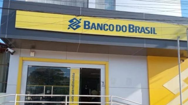 Fachada de uma agência do Banco do Brasil, com letreiro em destaque na parte superior, nas cores amarelo e azul. A entrada possui portas de vidro e rampa de acessibilidade com corrimão branco.