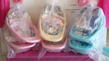 Seis kits maternidade embalados em plástico transparente, contendo banheiras e itens de bebê, como fraldas, roupas e produtos de higiene. Os kits estão dispostos sobre uma mesa coberta com tecido rosa, prontos para entrega às gestantes atendidas pelo programa Kit Natalidade da Prefeitura de Correntina.