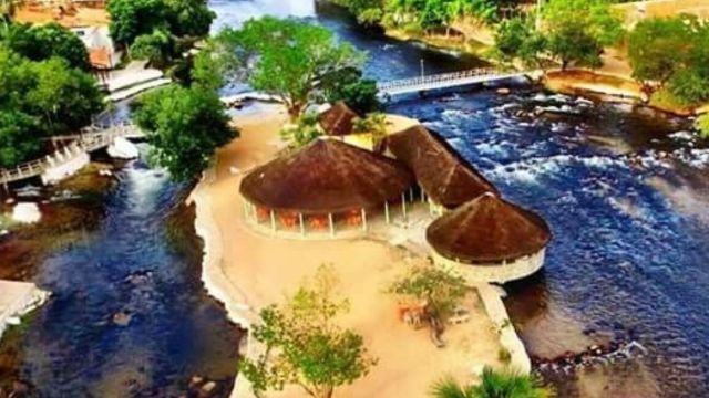 Vista aérea do Ranchão de Correntina, ponto turístico localizado no Rio Correntina, na Bahia. A foto mostra as construções cobertas por telhados de palha, cercadas por águas claras e árvores, com uma pequena ponte ligando as margens. O cenário natural destaca a beleza do local, que é um dos cartões-postais do município.