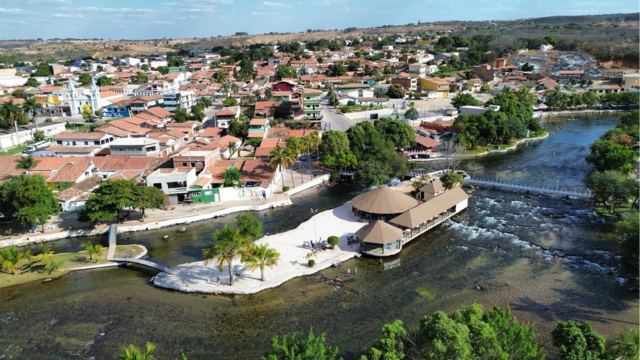 A imagem mostra uma vista aérea de Correntina, destacando o Ranchão construído sobre o rio de águas claras. O cenário reúne áreas de lazer, pequenas praias e vegetação ao redor do cartão-postal. Ao fundo, aparece o centro da cidade com casas, comércio e pontos históricos.