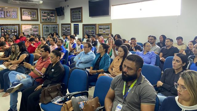 A imagem mostra uma sala cheia de participantes sentados em cadeiras azuis, assistindo atentamente ao 1º Fórum Comunitário do Selo UNICEF. O público é composto por homens e mulheres de diferentes idades, todos concentrados nas apresentações. As paredes ao fundo exibem diversos quadros e painéis, e a iluminação natural entra pela janela, contribuindo para um ambiente claro e organizado. O clima geral transmite engajamento e interesse nas discussões propostas pelo evento.