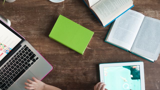 Mesa de estudos com livros abertos, um caderno verde fechado, um notebook e um tablet em uso, representando o uso combinado de materiais impressos e digitais na educação.