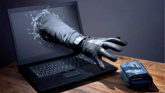 Ilustração de um braço com luva preta saindo de dentro de um laptop, como se estivesse quebrando a tela para tentar pegar uma carteira que está sobre a mesa, representando o conceito de crime virtual ou roubo digital.