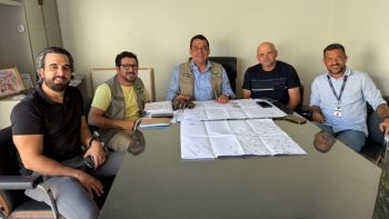 O prefeito Mariano Correntina, ao centro, participa de reunião com membros da equipe técnica. Todos estão sentados ao redor de uma mesa, analisando plantas e documentos referentes a obras no município. A imagem registra um momento de planejamento e alinhamento entre os gestores.
