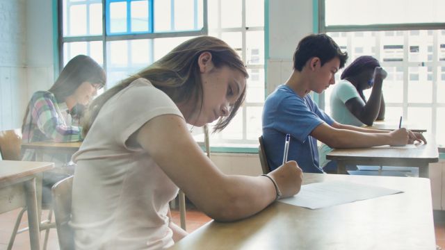 A imagem mostra um grupo de estudantes sentados em uma sala de aula, concentrados na realização de uma prova escrita. Em primeiro plano, uma aluna inclina levemente o corpo sobre a carteira enquanto escreve, demonstrando atenção e foco. Ao fundo, outros alunos também estão sentados individualmente, com canetas nas mãos, preenchendo folhas de papel. O ambiente é bem iluminado, com luz natural entrando pelas grandes janelas laterais, o que contribui para uma atmosfera clara e tranquila. As carteiras estão organizadas em fileiras, sugerindo um momento formal de avaliação, como um exame ou processo seletivo. A expressão corporal dos estudantes transmite silêncio, concentração e seriedade, características típicas de um período de prova escolar ou acadêmica.