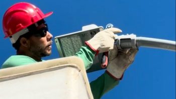 Na imagem, um trabalhador usa capacete de segurança vermelho, óculos escuros e luvas grossas enquanto instala uma luminária de LED em um poste de iluminação pública. Ele está em uma plataforma elevada contra o fundo de céu azul.