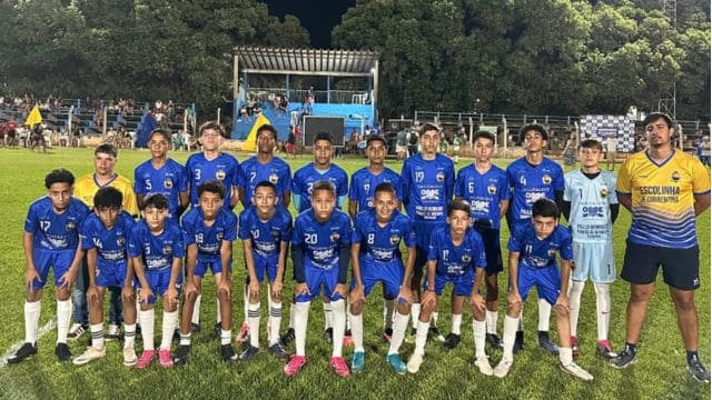 Um grupo de jovens atletas de futebol posa para foto em campo gramado à noite. Eles vestem uniforme azul com detalhes brancos, meias brancas e chuteiras coloridas. À direita, um treinador em uniforme amarelo e azul acompanha a equipe. Ao fundo, arquibancada com torcedores e árvores iluminadas completam o cenário.
