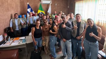 Prefeito Mariano em reunião com agricultores do PNAE e representantes da Prefeitura de Correntina em sala de reuniões, com bandeiras do Brasil e do município ao fundo. Pessoas em pé, olhando para a câmera e fazendo gesto de positivo com o polegar, simbolizando união e parceria.
