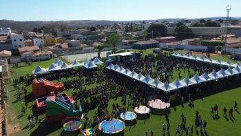 Imagem aérea mostra uma grande estrutura montada em um campo gramado, com dezenas de tendas brancas enfileiradas formando corredores. Uma multidão de pessoas — crianças, pais e educadores — circula pelo local. No canto esquerdo, brinquedos infláveis e camas elásticas coloridas são utilizados pelas crianças. Ao fundo, é possível ver casas e prédios residenciais da cidade. A cena representa um evento público festivo, organizado pela Prefeitura de Correntina, em alusão à entrega de uniformes e material escolar.