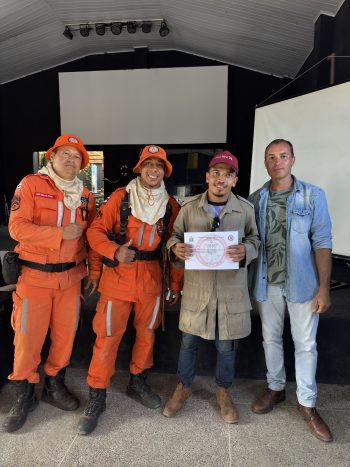 Formando do curso de brigadista voluntário de Correntina posa com certificado ao lado de dois instrutores do Corpo de Bombeiros de Barreiras, ambos uniformizados em trajes laranja de combate a incêndios, e dois representantes da organização. A foto foi tirada no encerramento da capacitação promovida pela Prefeitura de Correntina, em ambiente interno, com fundo escuro e tela de projeção ao fundo. Todos os presentes demonstram orgulho e satisfação com a conquista.