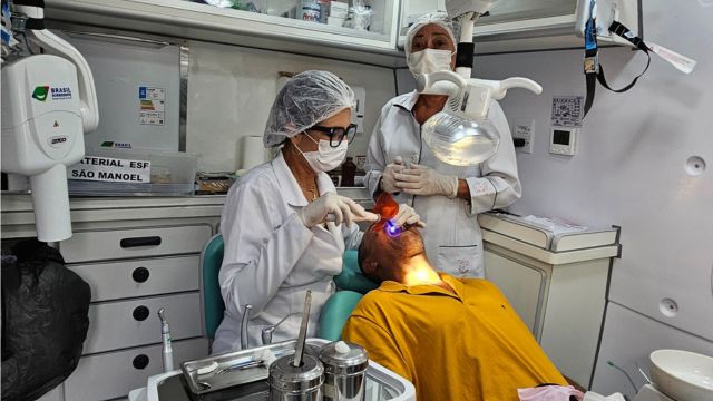 Profissionais de saúde realizam atendimento odontológico em um consultório móvel. Uma dentista, usando jaleco branco, touca e máscara, atende um paciente de camiseta amarela, deitado na cadeira odontológica. Outro profissional acompanha o procedimento ao fundo, também paramentado com equipamentos de proteção