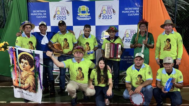 Grupo de homens e uma mulher vestindo camisetas verdes participa de evento cultural em Correntina, segurando instrumentos musicais como sanfona, pandeiro e triângulo, em frente a um painel com a identidade visual do “Encontro de Reisado 2025” e da Secretaria Municipal de Cultura.