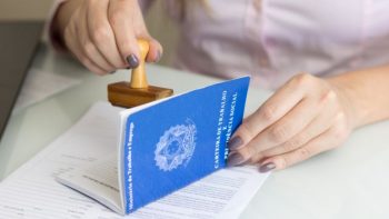 Uma pessoa carimbando uma Carteira de Trabalho e Previdência Social (CTPS) azul sobre uma mesa branca, simbolizando a formalização de um emprego.