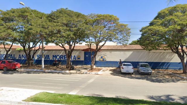 Imagem da fachada de uma escola municipal, com prédio térreo pintado nas cores azul e branco, árvores grandes em frente ao muro, alguns carros estacionados ao longo da via e uma rua asfaltada em primeiro plano, em um dia ensolarado.