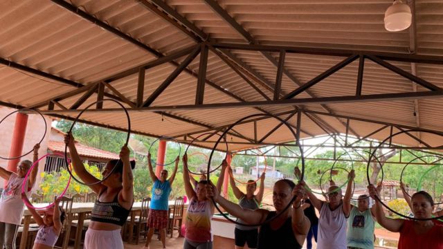 Grupo de mulheres de diversas idades participa de uma atividade física em um espaço coberto. Elas aparecem de pé, dispostas em filas, segurando bambolês pretos acima da cabeça com as duas mãos. O local possui estrutura de telhado metálico aparente e pilares vermelhos.