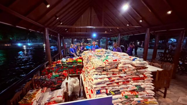 Fotografia noturna em plano médio mostrando uma grande quantidade de alimentos arrecadados dispostos em mesas dentro de um quiosque ou píer de madeira com teto rústico e iluminação quente. No primeiro plano e ao centro, destaca-se uma enorme pilha organizada de pacotes de arroz e feijão ensacados. À esquerda, há outra bancada repleta de alimentos em embalagens coloridas menores e fardos variados. Ao fundo, um grupo de cerca de sete pessoas veste camisetas azuis e roxas (aparentemente a equipe de organização do evento), conversando atrás das mesas de alimentos. À esquerda da estrutura, é possível ver as águas escuras de um rio refletindo as luzes urbanas da noite. À direita, mesas e cadeiras de madeira completam o ambiente interno do quiosque.