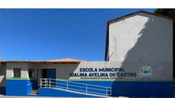 Fotografia da fachada da Escola Municipal Idalina Avelina de Castro. O edifício térreo possui paredes pintadas de cinza-claro na parte superior e azul na base, com uma faixa verde a separar as cores. No centro, destaca-se o nome da escola e o brasão do município de Correntina em letras azuis e amarelas. À esquerda, há uma rampa de acessibilidade com corrimão branco que dá acesso à entrada principal. O dia está ensolarado e o céu está completamente limpo e azul.