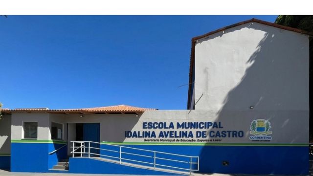 Fotografia da fachada da Escola Municipal Idalina Avelina de Castro. O edifício térreo possui paredes pintadas de cinza-claro na parte superior e azul na base, com uma faixa verde a separar as cores. No centro, destaca-se o nome da escola e o brasão do município de Correntina em letras azuis e amarelas. À esquerda, há uma rampa de acessibilidade com corrimão branco que dá acesso à entrada principal. O dia está ensolarado e o céu está completamente limpo e azul.