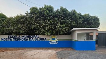 Fotografia da fachada e do muro da Escola Municipal Nossa Senhora da Glória. O muro possui a parte superior pintada de branco e a base de azul, com uma fina listra verde a dividir as cores. À esquerda, sob a copa de árvores densas, destaca-se a inscrição