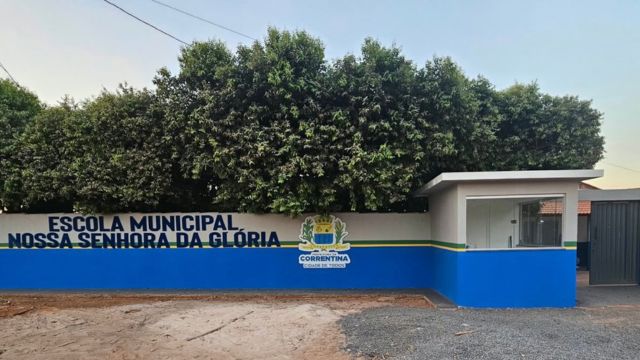 Fotografia da fachada e do muro da Escola Municipal Nossa Senhora da Glória. O muro possui a parte superior pintada de branco e a base de azul, com uma fina listra verde a dividir as cores. À esquerda, sob a copa de árvores densas, destaca-se a inscrição "ESCOLA MUNICIPAL NOSSA SENHORA DA GLÓRIA" acompanhada pelo brasão da Prefeitura de Correntina. À direita, encontra-se uma guarita de segurança com janelas de vidro e, ao lado, um portão cinza de acesso. O pátio em frente combina uma área de terra batida com uma secção coberta por brita cinzenta.