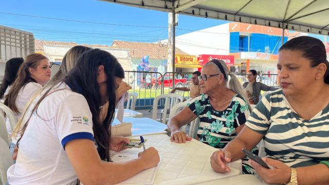 Profissional da Secretaria Municipal de Saúde atende moradoras presencialmente em uma mesa sob tenda, preenchendo fichas de cadastro durante o mutirão de saúde.