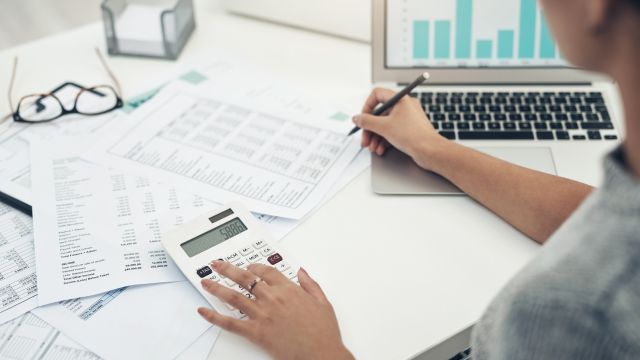 Vista superior parcial de uma mesa de trabalho organizada. Uma mão feminina digita na calculadora enquanto a outra segura uma caneta sobre documentos financeiros. Óculos de grau e um notebook com gráficos completam a cena de análise técnica.