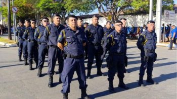 Fotografia em plano médio de um grupo de aproximadamente dez guardas civis municipais perfilados em uma rua ao ar livre, durante o dia. Eles vestem uniformes operacionais na cor azul-marinho com detalhes em amarelo nos ombros, boinas pretas e cinturões de utilidades. Os guardas estão em posição de descanso, com as mãos para trás, organizados em duas fileiras. Ao fundo, é possível observar árvores, uma calçada e uma placa de sinalização de trânsito indicando limite de velocidade de 30 km/h.