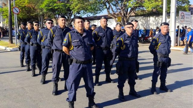 Fotografia em plano médio de um grupo de aproximadamente dez guardas civis municipais perfilados em uma rua ao ar livre, durante o dia. Eles vestem uniformes operacionais na cor azul-marinho com detalhes em amarelo nos ombros, boinas pretas e cinturões de utilidades. Os guardas estão em posição de descanso, com as mãos para trás, organizados em duas fileiras. Ao fundo, é possível observar árvores, uma calçada e uma placa de sinalização de trânsito indicando limite de velocidade de 30 km/h.