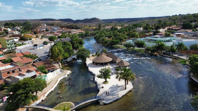 Vista aérea do Ranchão da Orla de Correntina, com o rio de águas cristalinas ao redor, área de convivência, quiosques, palmeiras e casas da cidade ao fundo, destacando um dos principais pontos turísticos do município.