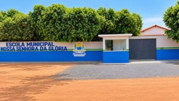 Fachada da Escola Municipal Nossa Senhora da Glória, em Correntina. O muro frontal está pintado de branco na parte superior e azul na parte inferior, com uma faixa verde e amarela ao centro. Do lado esquerdo, sobressai o nome da escola em letras azuis e o brasão da prefeitura de Correntina. Ao lado, há uma guarita de acesso pintada nas mesmas cores e um portão de garagem cinza. Ao fundo, veem-se árvores copadas e verdes sob um céu límpido.