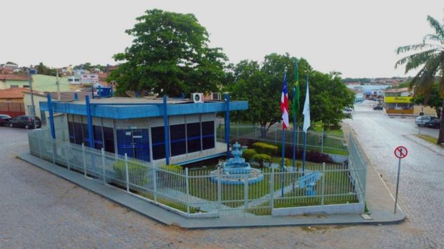 Fachada da sede do IMUPRE, em Correntina, com jardim, fonte e bandeiras na área externa.
