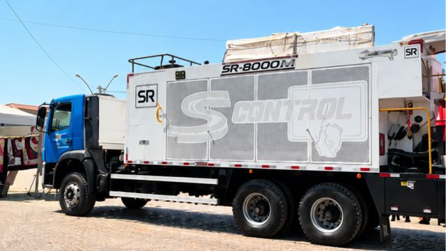 Caminhão azul equipado com usina de micropavimentação asfáltica, estacionado em área pavimentada. O equipamento possui estrutura branca e cinza, com as inscrições “SR-8000M” e “S Control” na lateral.