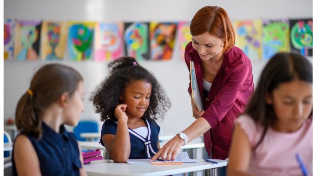 Professora orienta aluna em sala de aula, apontando atividade sobre a mesa, enquanto outras crianças participam da aula ao fundo, com trabalhos coloridos expostos na parede.