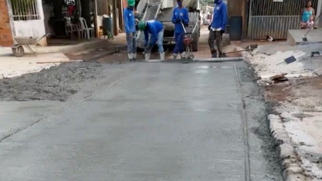 Quatro trabalhadores da Prefeitura, utilizando uniformes azuis e equipamentos de proteção, realizam o nivelamento de uma camada de concreto recém-aplicada em uma via urbana. Ao fundo, um caminhão betoneira fornece o material, enquanto os profissionais utilizam uma régua vibratória para dar acabamento ao pavimento. A cena registra uma obra de infraestrutura e pavimentação em uma rua residencial.