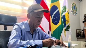 Homem de camisa azul e boné cinza escreve em um caderno sobre a mesa de um gabinete, com bandeiras do Brasil, da Bahia e do município ao fundo.