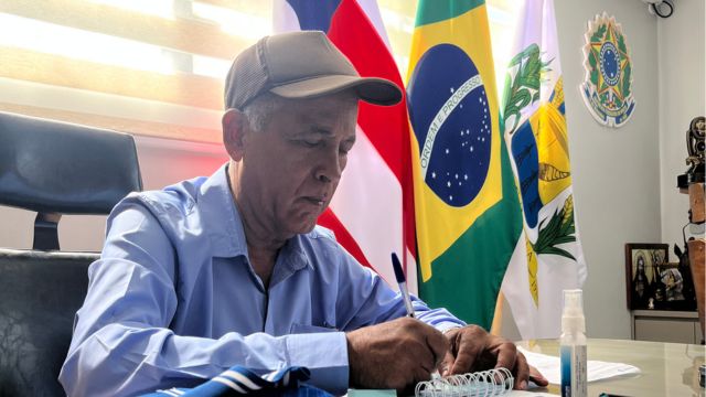 Homem de camisa azul e boné cinza escreve em um caderno sobre a mesa de um gabinete, com bandeiras do Brasil, da Bahia e do município ao fundo.