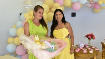 Gestante de vestido verde segura um kit natalidade com banheira e produtos para os cuidados com o bebê, ao lado de uma mulher de vestido amarelo, durante entrega em espaço decorado com balões coloridos.