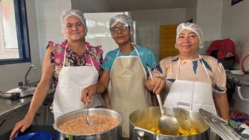 Três profissionais da alimentação escolar, usando toucas e aventais, posam para foto na cozinha de uma escola, ao lado de panelas com alimentos preparados para os estudantes.