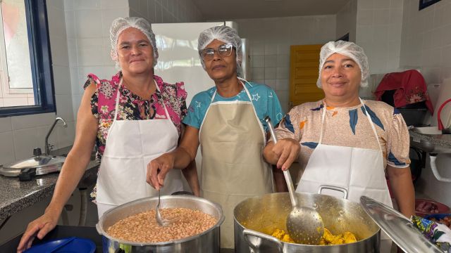 Três profissionais da alimentação escolar, usando toucas e aventais, posam para foto na cozinha de uma escola, ao lado de panelas com alimentos preparados para os estudantes.