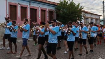 Integrantes da Filarmônica Erato-Correntinense marcham pelas ruas de Correntina tocando instrumentos de sopro, como trompetes, trombones e saxofones, durante uma apresentação pública. Os músicos vestem camisetas azuis e seguem em desfile por uma rua de paralelepípedo, acompanhados por pessoas ao redor, representando a tradição musical e cultural do município.