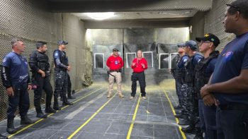 Grupo de agentes da Guarda Civil Municipal em treinamento de tiro em estande fechado, organizados em duas fileiras, enquanto dois instrutores ao centro orientam a atividade diante de alvos posicionados ao fundo.