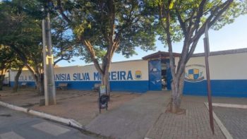 Fotografia da fachada da Escola Municipal Anísia Silva Moreira sob a luz do dia. O prédio possui um muro branco com a metade inferior pintada de azul, onde se lê o nome da instituição em letras grandes e azuis. À direita, há um portão de metal azul e o brasão da escola estampado na parede. O espaço à frente é composto por uma calçada de blocos intertravados, árvores de copa densa que projetam sombras no chão e bancos de praça vazios. A área externa está limpa e sem a presença de pessoas.