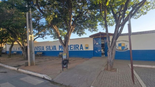 Fotografia da fachada da Escola Municipal Anísia Silva Moreira sob a luz do dia. O prédio possui um muro branco com a metade inferior pintada de azul, onde se lê o nome da instituição em letras grandes e azuis. À direita, há um portão de metal azul e o brasão da escola estampado na parede. O espaço à frente é composto por uma calçada de blocos intertravados, árvores de copa densa que projetam sombras no chão e bancos de praça vazios. A área externa está limpa e sem a presença de pessoas.