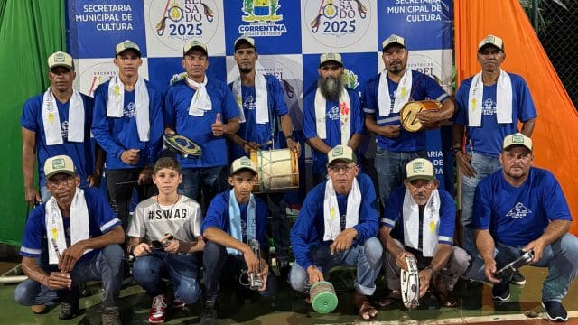 Grupo de músicos com camisetas azuis posa para foto, alguns segurando instrumentos musicais, diante de um painel da Secretaria Municipal de Cultura de Correntina, durante evento cultural em 2025.
