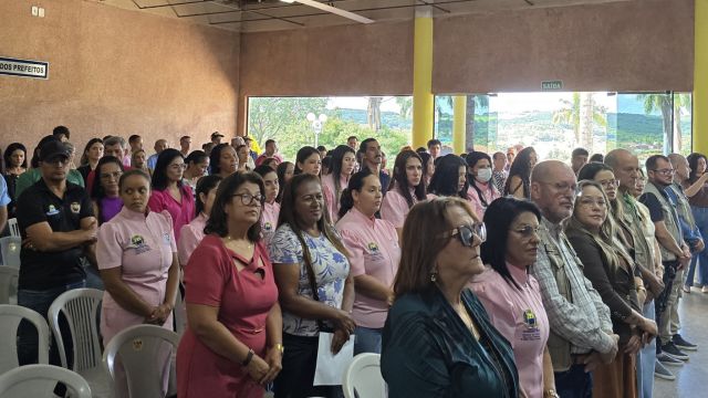 Grupo de pessoas reunidas em um espaço fechado, em pé, organizadas em fileiras, participando de um evento ou cerimônia. Muitas usam camisetas rosa com identificação, enquanto outras vestem roupas variadas. O ambiente parece ser um auditório ou salão comunitário, com cadeiras ao fundo e paredes com pinturas. A maioria das pessoas está voltada para a mesma direção, demonstrando atenção a alguma atividade ou fala.