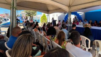 Participantes acompanham uma solenidade realizada sob uma tenda, com cadeiras ocupadas pelo público voltadas para uma mesa de autoridades. Ao fundo, um púlpito é utilizado para pronunciamentos, enquanto um painel azul com balões decora o espaço. O evento acontece durante o dia, em área externa, com céu limpo e presença de autoridades e convidados.