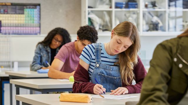 A imagem mostra uma sala de aula durante a aplicação de uma prova de concurso. Em primeiro plano, uma estudante jovem está sentada em uma carteira individual, concentrada enquanto escreve em uma folha de papel. Ela usa uma camiseta listrada sob um macacão jeans e mantém postura inclinada sobre a mesa, demonstrando foco na tarefa. Ao fundo, outros candidatos também realizam a prova, sentados em fileiras organizadas. O ambiente é claro, bem iluminado e estruturado, com estantes contendo livros e materiais didáticos, além de um quadro ou painel com conteúdos afixados na parede. A cena transmite um clima de atenção, organização e dedicação aos estudos, sugerindo um momento de avaliação individual em contexto de concurso público.