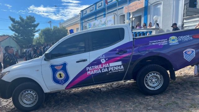 Viatura branca e roxa da Patrulha Maria da Penha estacionada em frente a um prédio, com pessoas ao fundo acompanhando o evento.