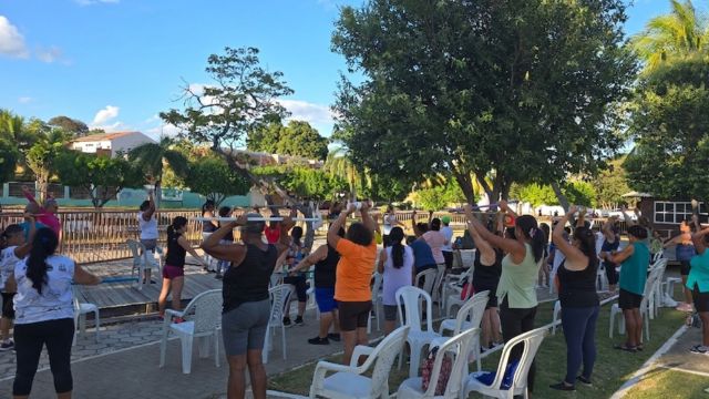 Uma fotografia ao ar livre mostra um grupo de pessoas, majoritariamente idosas, participando de uma aula de ginástica em um espaço público arborizado. Os participantes estão de pé, dispostos em fileiras, e realizam um exercício de levantamento de bastões acima da cabeça. Várias cadeiras brancas de plástico estão espalhadas pelo local, servindo de apoio entre os alunos. O ambiente é ensolarado, com gramado, árvores densas ao fundo e parte de uma estrutura de madeira à esquerda.